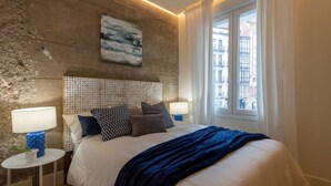 Ein modernes Schlafzimmer mit einem großen Bett, einem strukturierten Kopfteil und Blick auf Gebäude durch ein Fenster.
