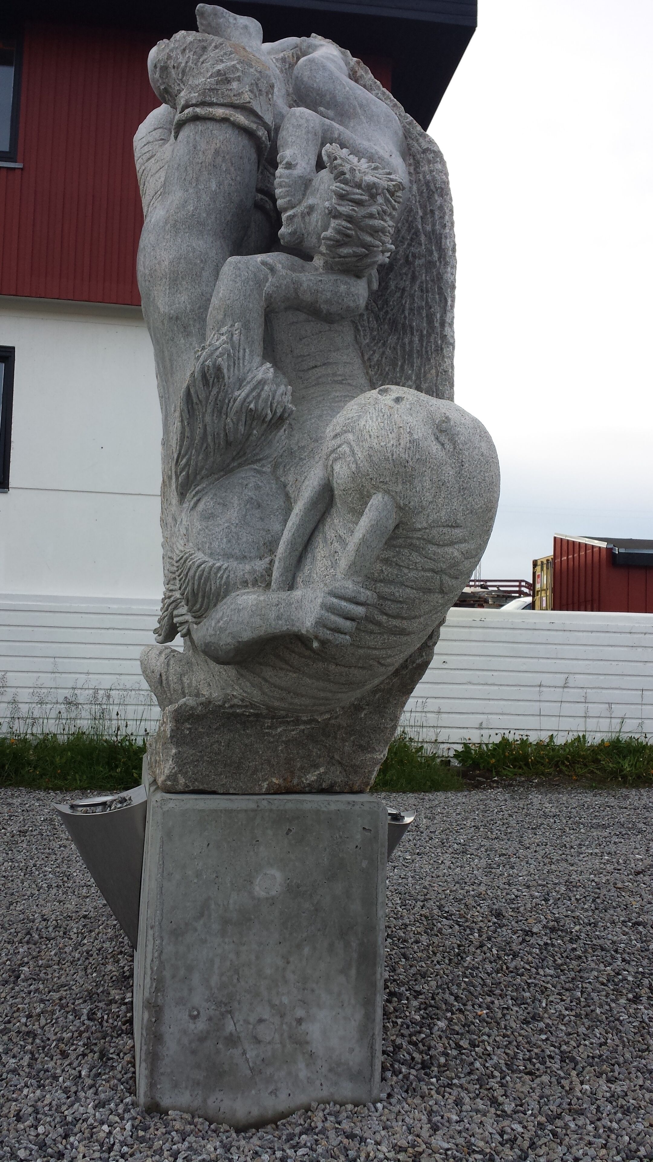 A stone sculpture of a reclining figure with a child, set on a concrete pedestal in an outdoor area with gravel ground and a red building in the background.