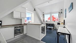 A compact kitchen with a built-in oven, a dining area with a table and chairs, and a view of red buildings through large windows. - Hotel Nordbo (Nuuk)