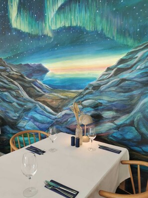 A dining table set with wine glasses, a vase with dried plants, and salt and pepper shakers, against a backdrop of a painting depicting a mountainous landscape under a starry sky. - Hotel Nordbo (Nuuk)