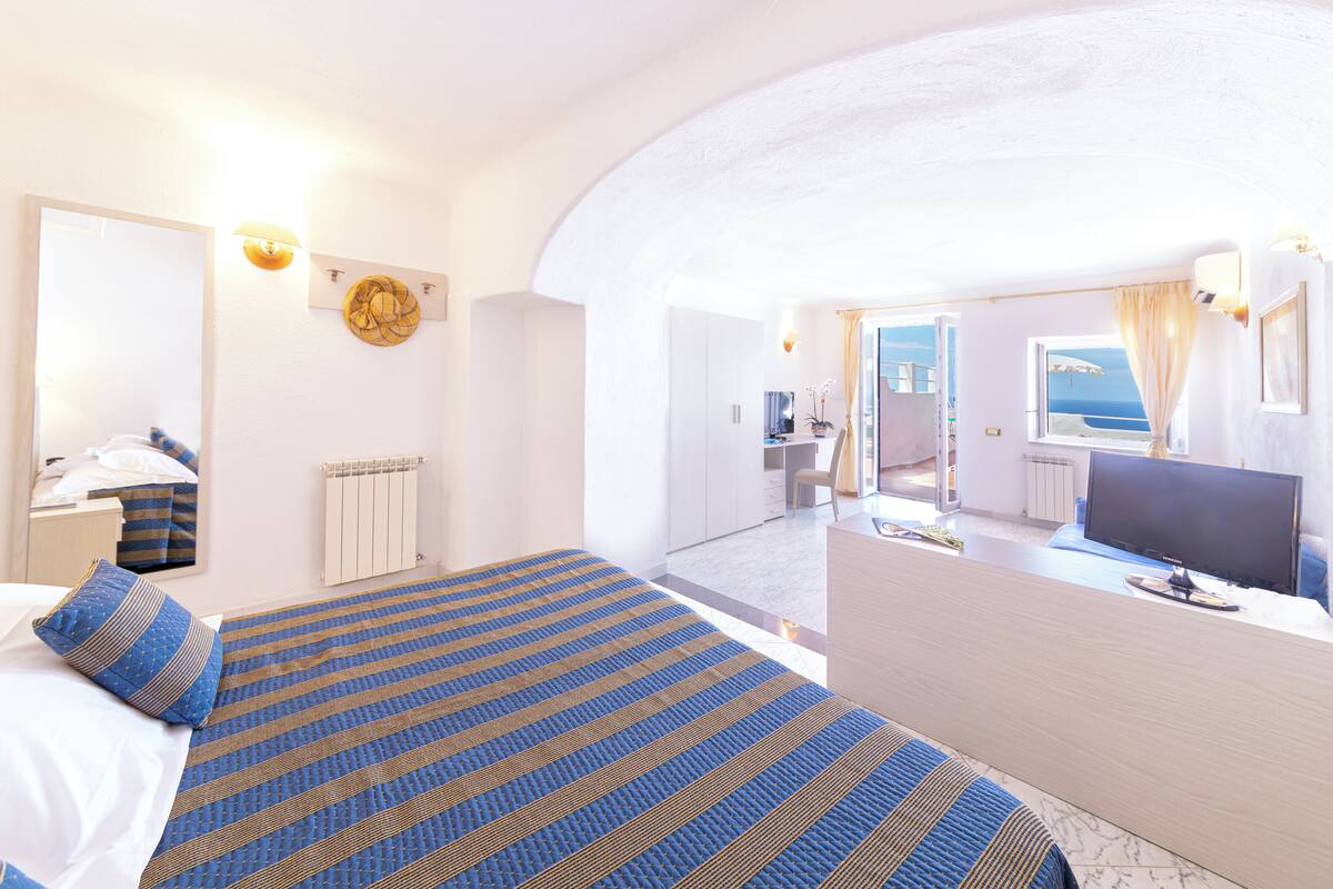 A hotel room with a bed, a desk, a TV, and a view of the sea.
