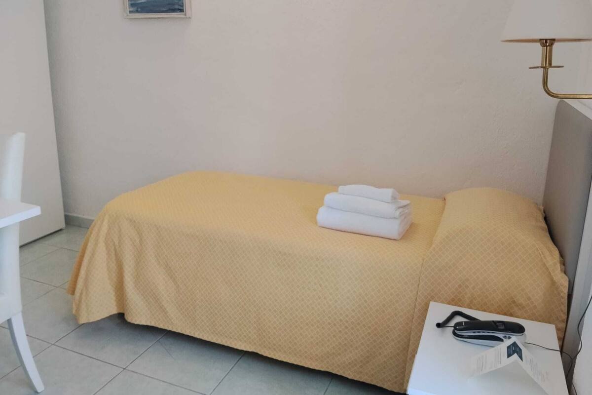 A small, single-bed room with a yellow bedspread, a white chair, a bedside table with a lamp, and a framed picture on the wall.