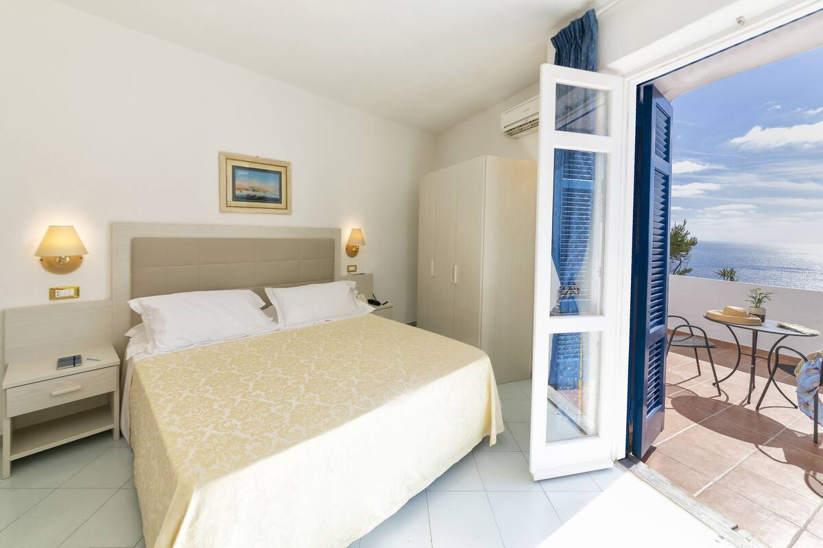 A hotel room with a bed, bedside lamps, a wardrobe, and a balcony with a view of the sea.