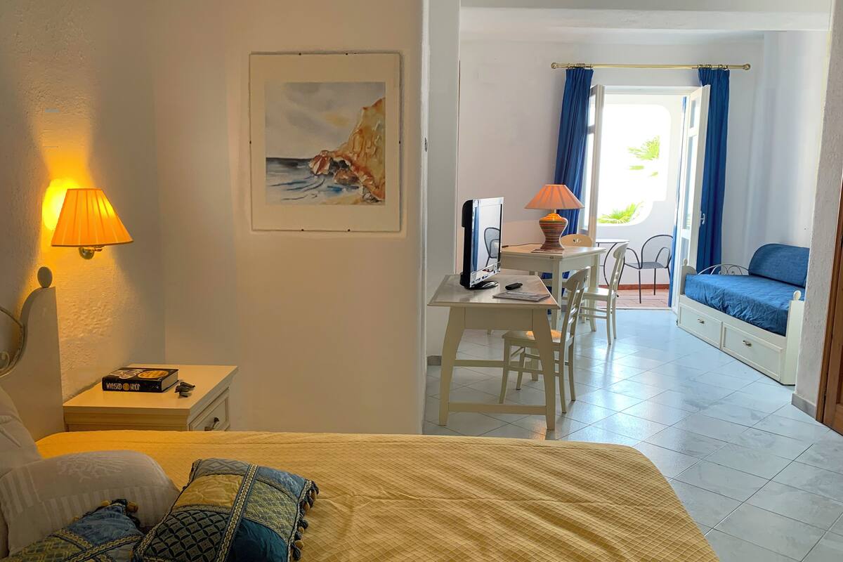 A hotel room with a bed, a desk with a computer, a chair, and a view of the sea through the window.
