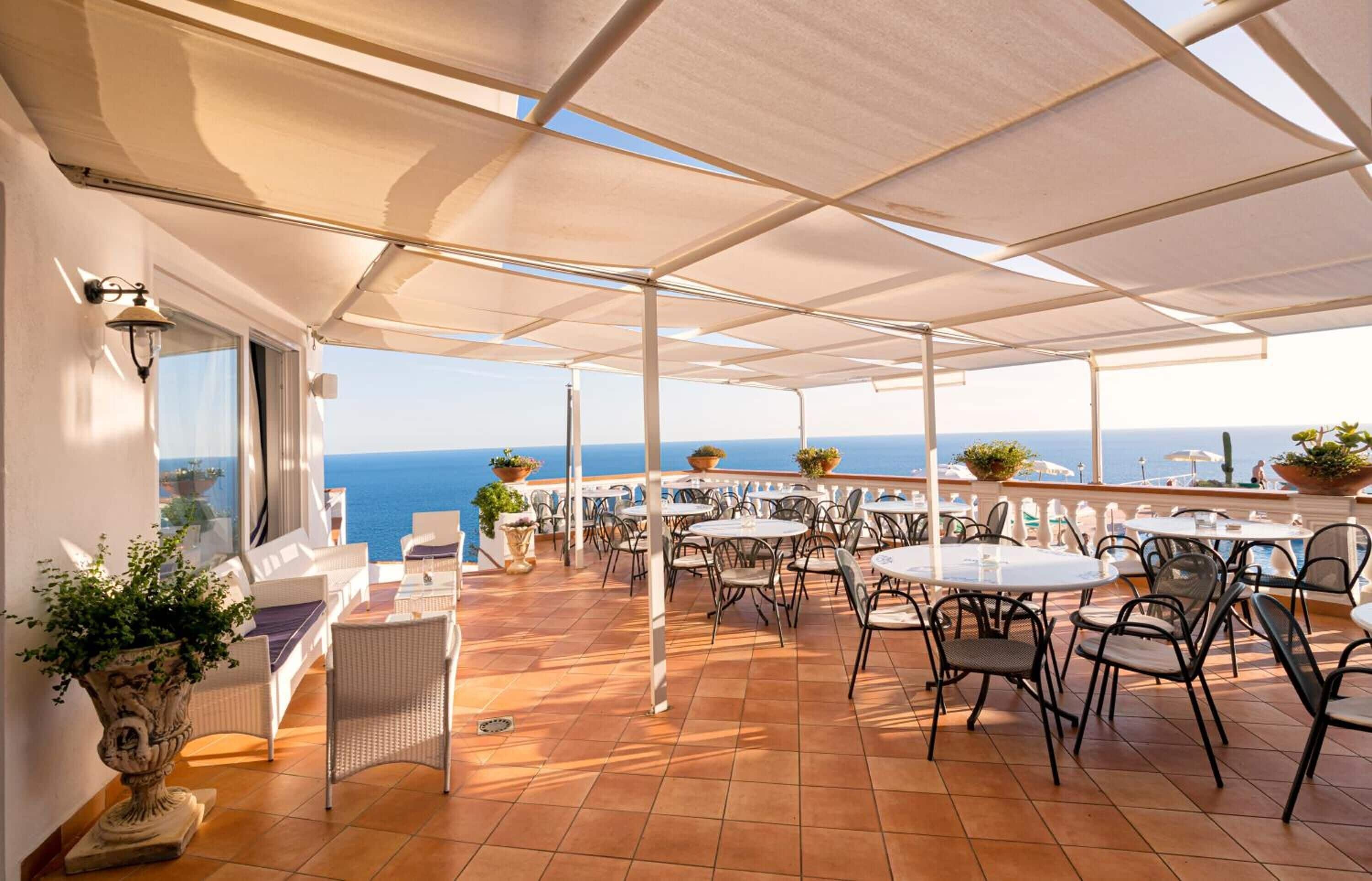 An outdoor terrace with multiple tables and chairs, potted plants, and a view of the sea.