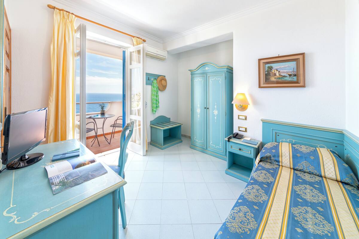 A room with a bed, a desk, a chair, a wardrobe, and a view of the sea.