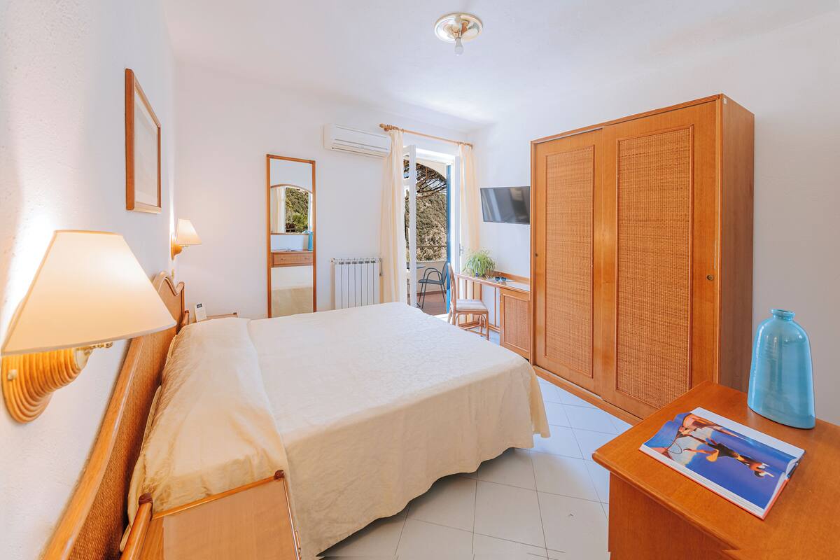 A bedroom with a bed, bedside lamps, a wardrobe, a small table with a magazine, and a view of the outside.