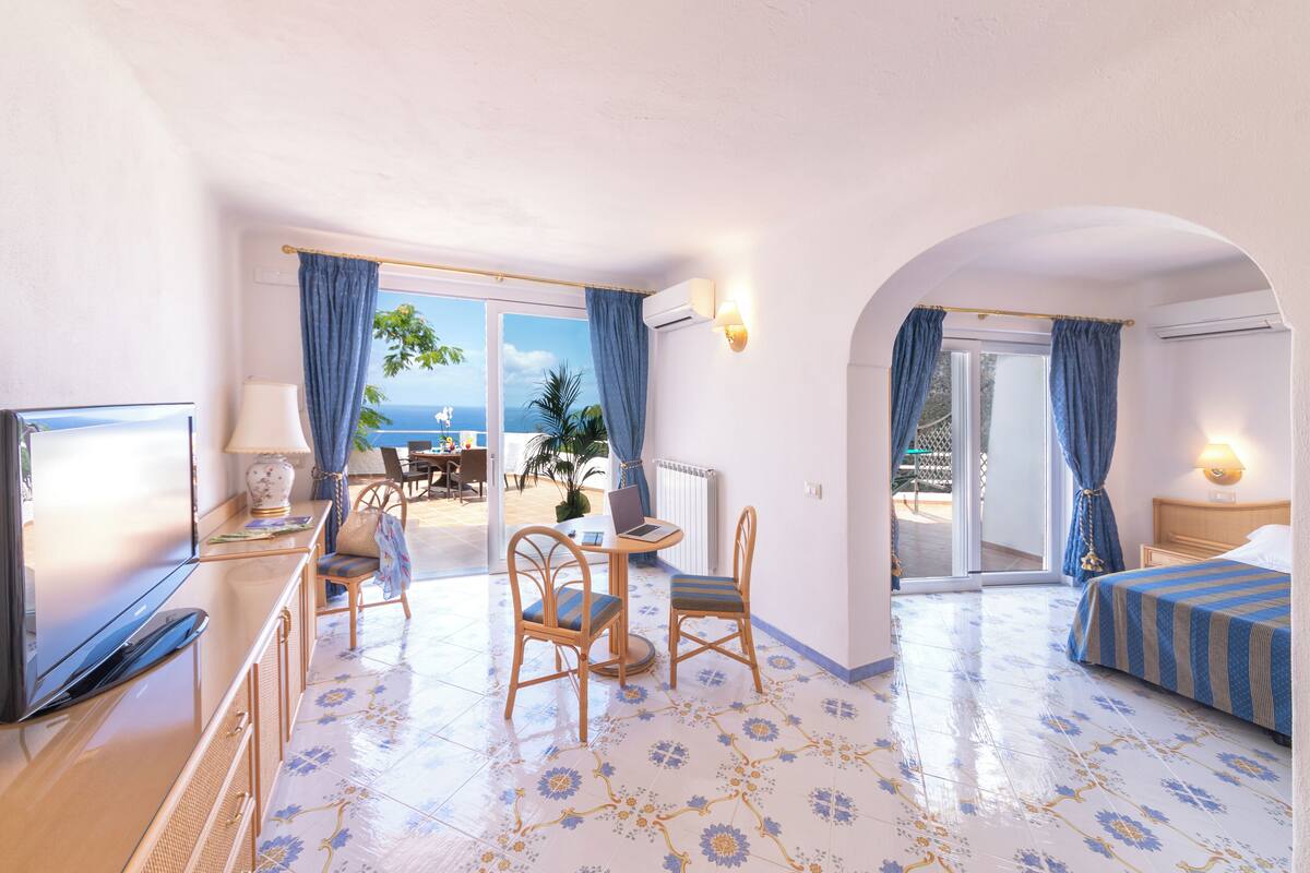 A hotel room with a bed, a desk with a laptop, a chair, and a view of the sea through large windows.