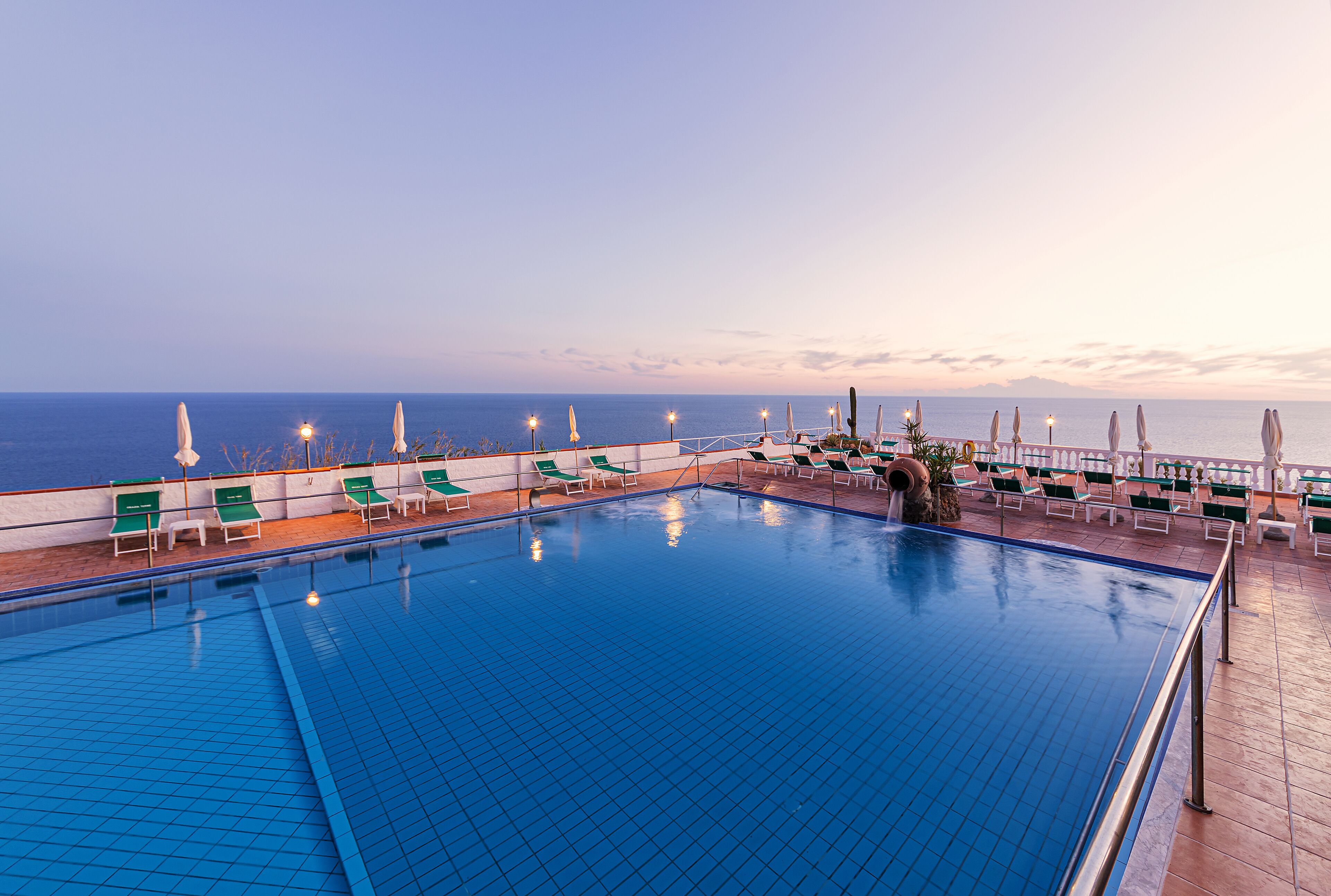 A swimming pool with green chairs and umbrellas, overlooking the sea.