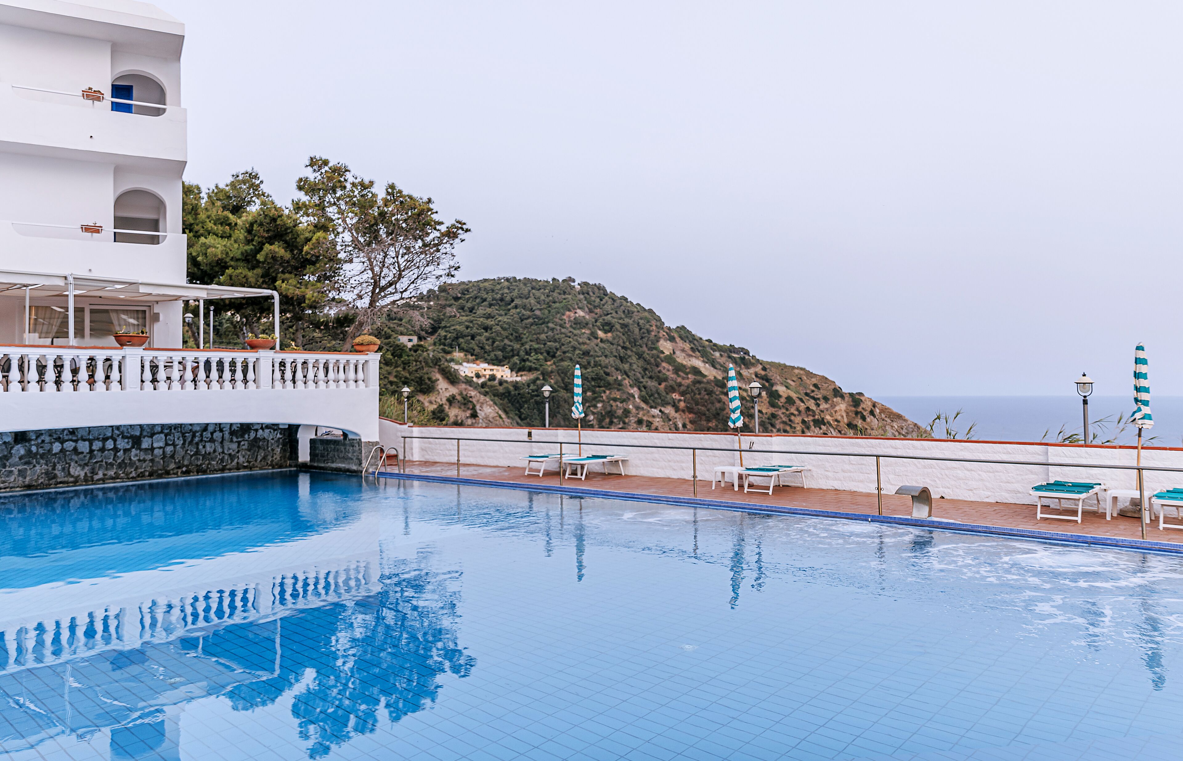 A swimming pool with a white building, a stone wall, and a view of a hill with trees and a body of water.