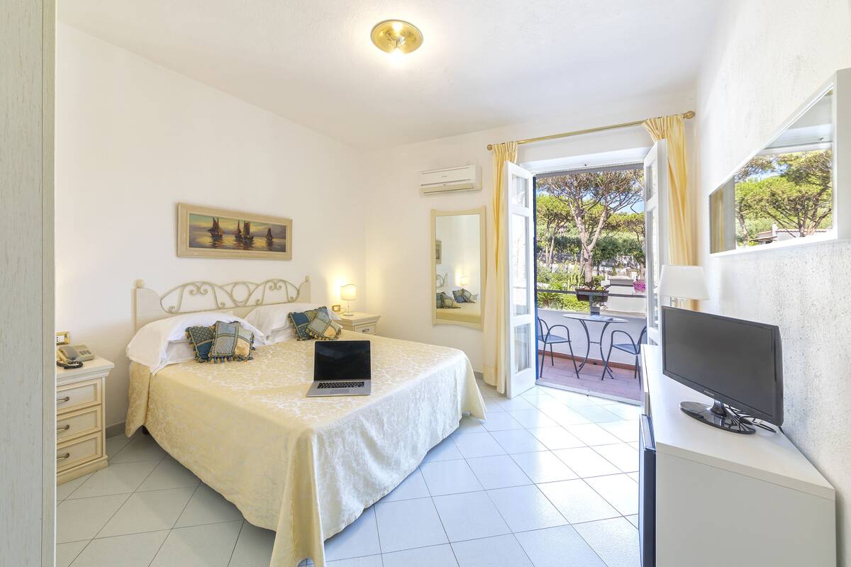 A bedroom with a bed, a laptop, a TV, and a balcony with outdoor furniture.