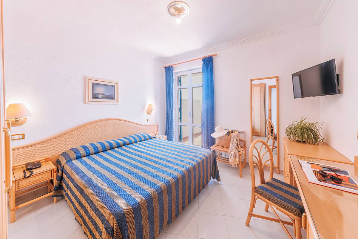 A hotel room with a bed, bedside table, lamp, desk, chair, TV, and a window with curtains.