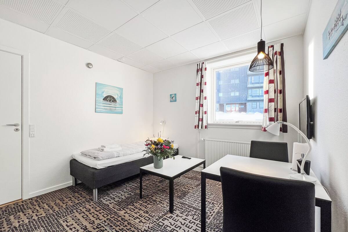 A hotel room with a bed, a desk, a chair, a small table, a vase with flowers, a window with curtains, a wall-mounted TV, and a ceiling with a light fixture.