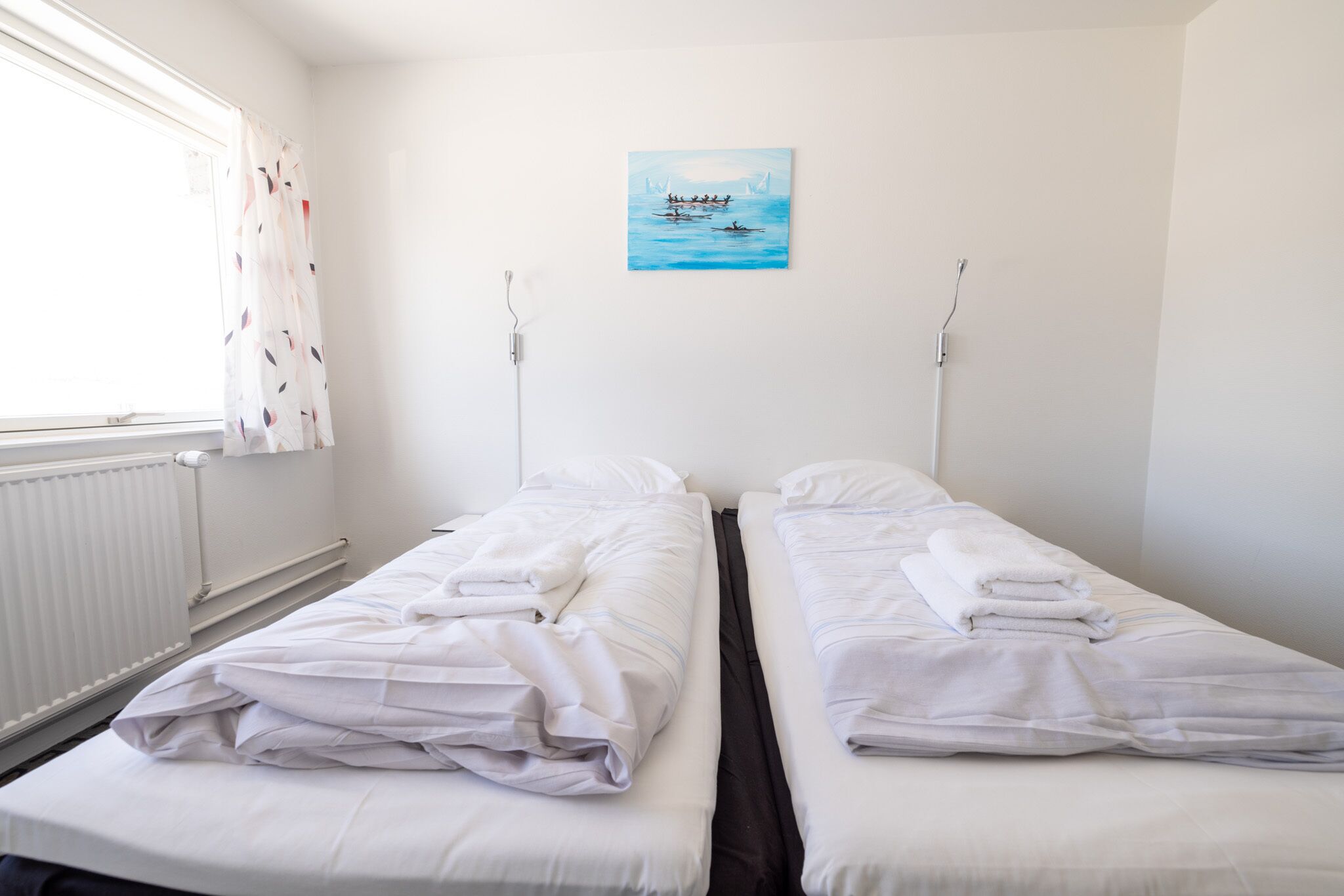 Habitación de hotel con dos camas individuales, ropa de cama blanca, una ventana con cortinas, un radiador y un cuadro de un barco en la pared.