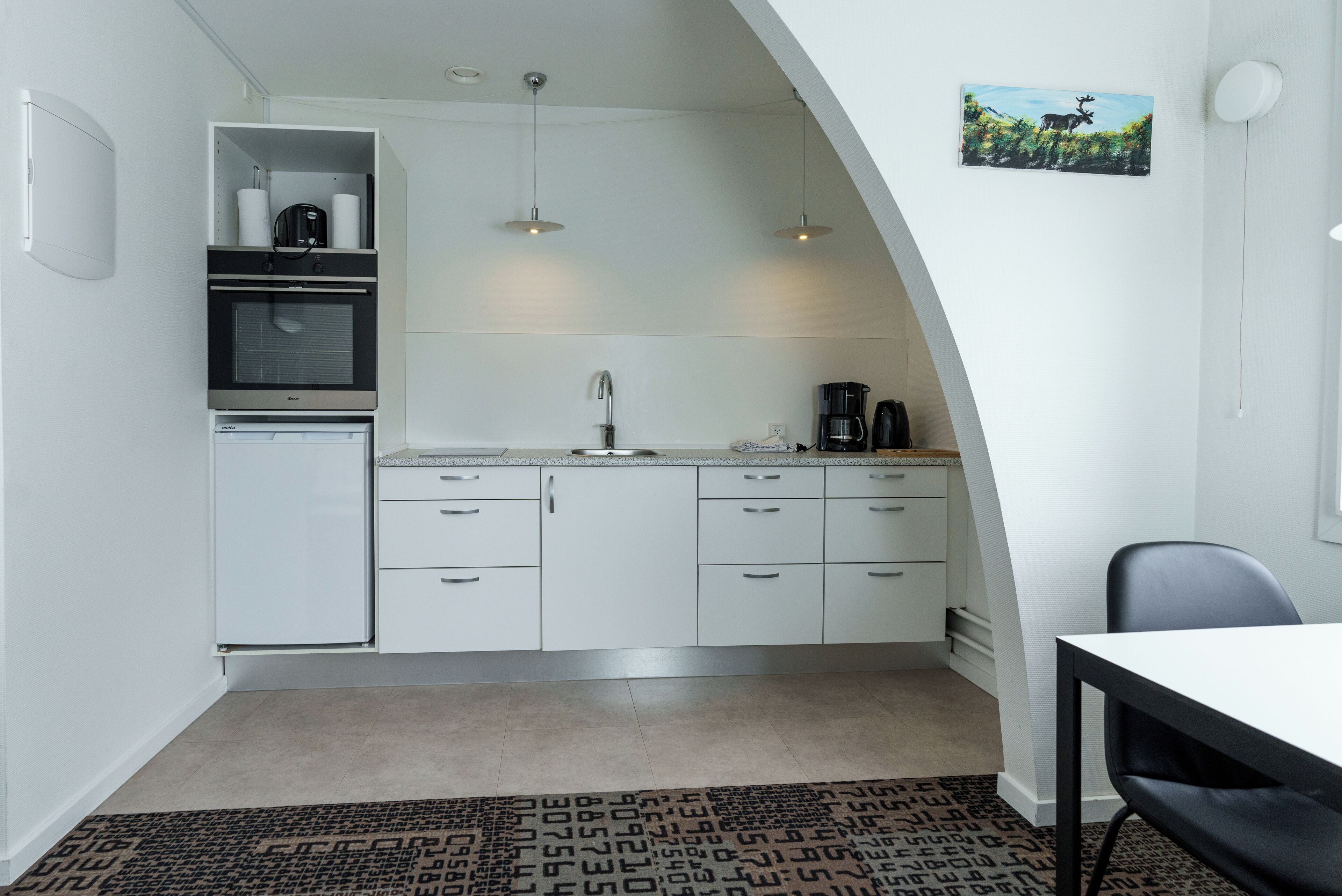 A compact kitchen with white cabinets, a built-in oven, a sink, and a coffee maker. There is a small dining area with a table and chairs.