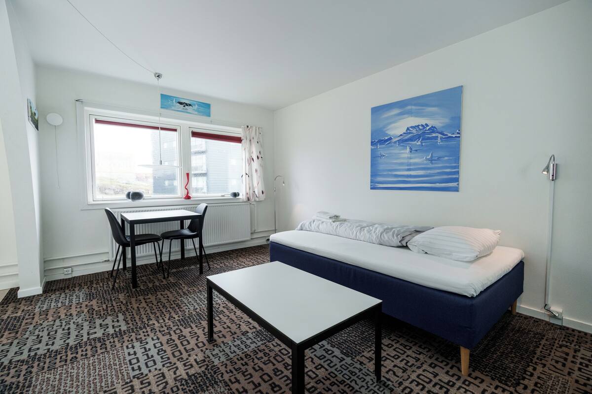 A room with a bed, a table, and a chair. There is a window with curtains and a painting on the wall.
