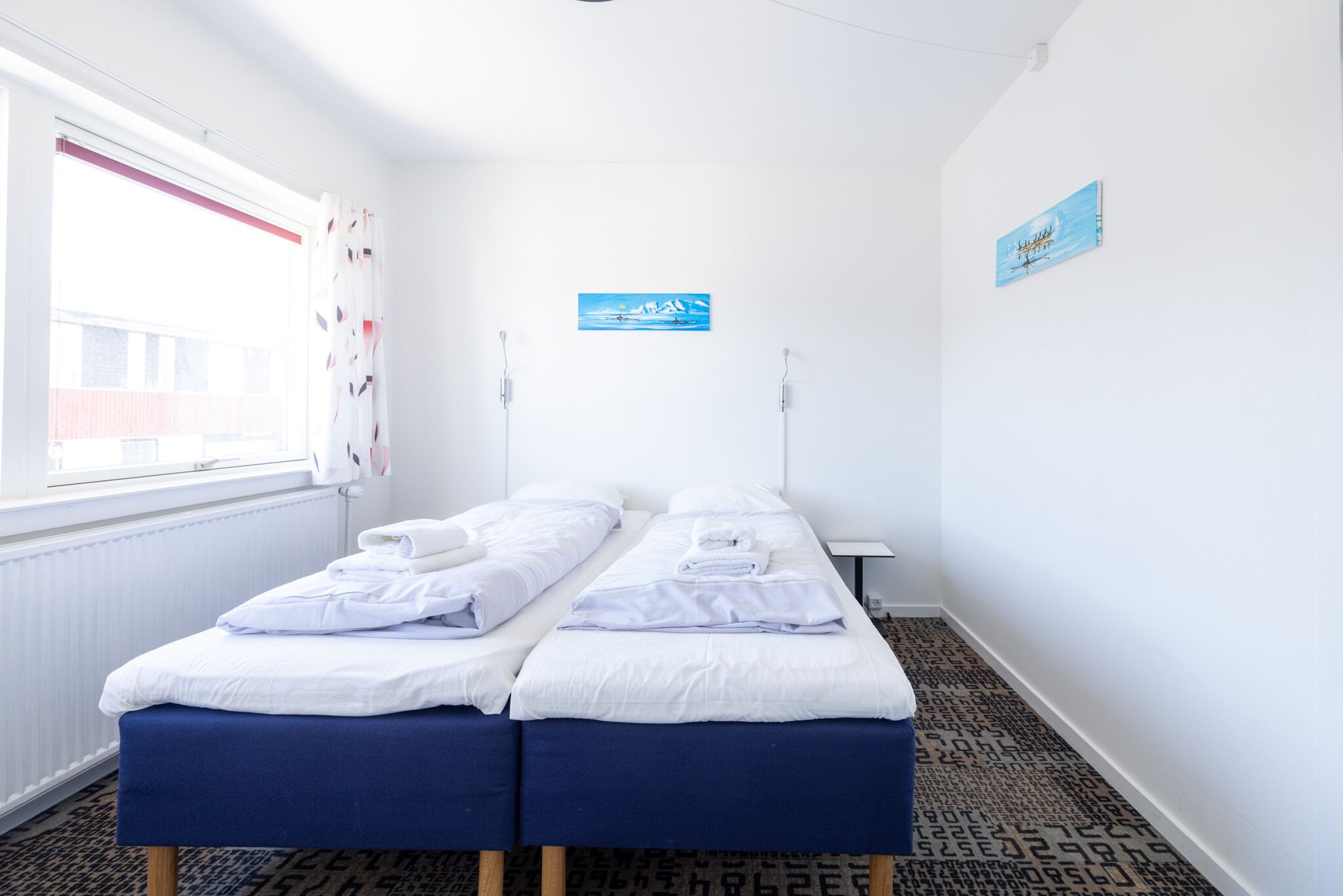 Een hotelkamer met twee enkele bedden, een raam met gordijnen, een radiator en een klein tafeltje.