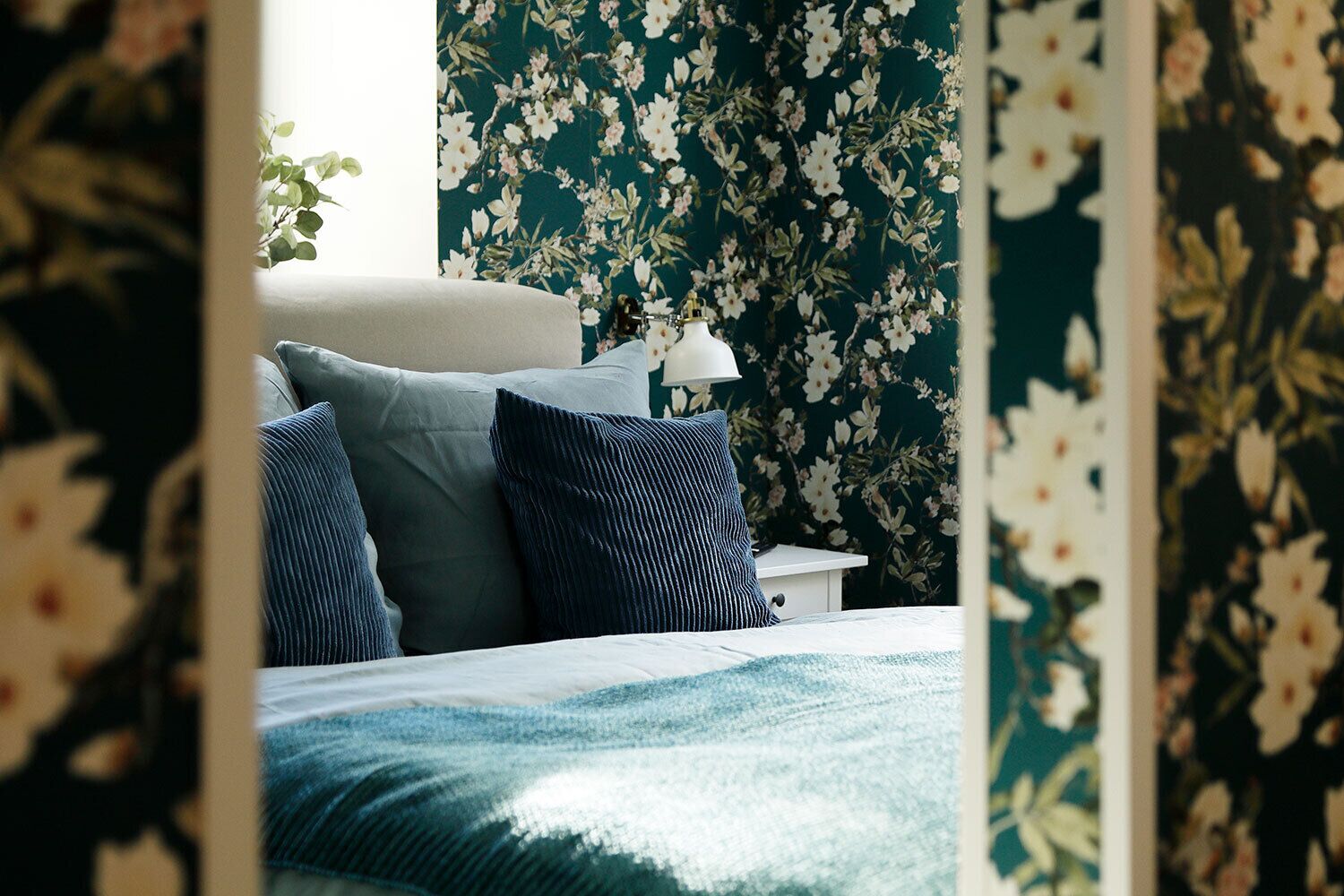A bedroom with a bed, pillows, a bedside table, a lamp, and floral wallpaper.