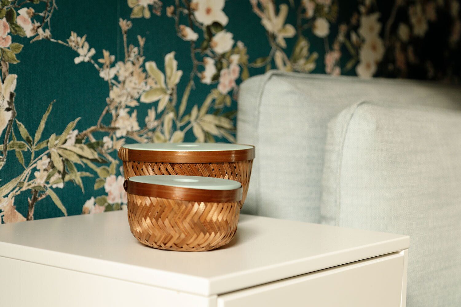 Two woven baskets on a white bedside table against a floral wallpaper background.