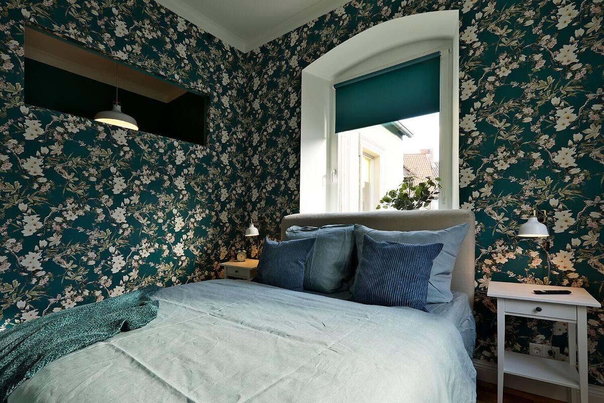 A bedroom with floral wallpaper, a bed with blue bedding, a nightstand with a lamp, and a window with a view of the outside.