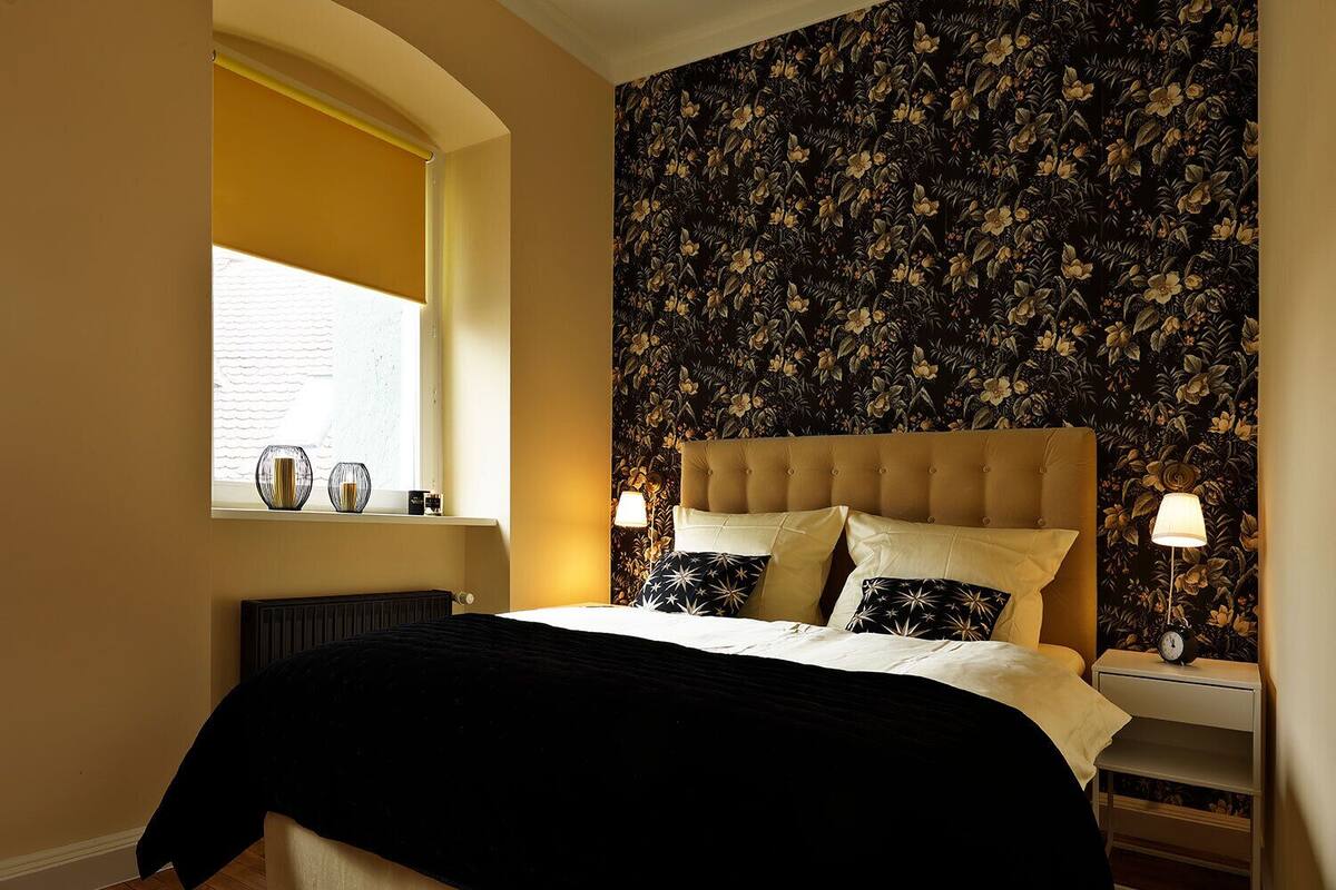 A bedroom with a large bed, two bedside tables, a window with a view, and a floral wallpaper.