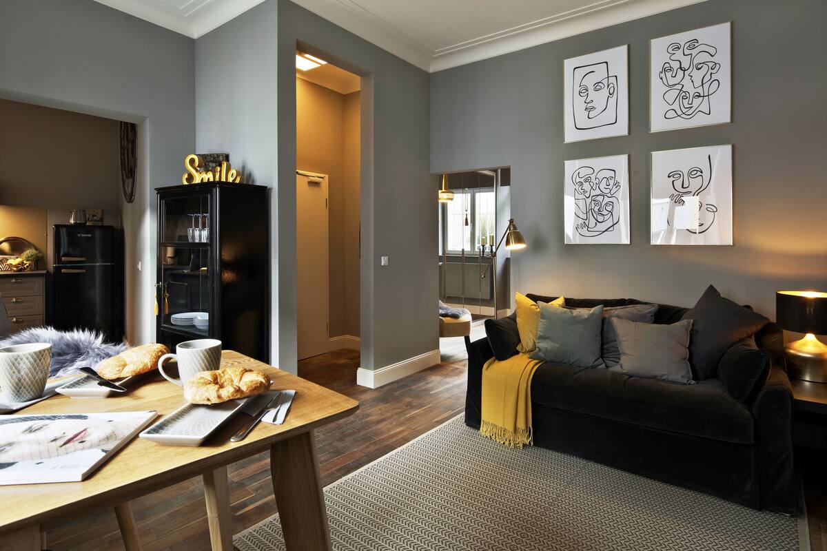 A modern living room with a dark sofa, a wooden dining table, and a kitchen area visible in the background.