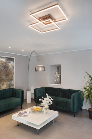 Una sala de estar moderna con un sofá verde, una mesa de centro blanca con un cuenco de frutas y flores, un cuadro abstracto grande y una lámpara de techo geométrica original.