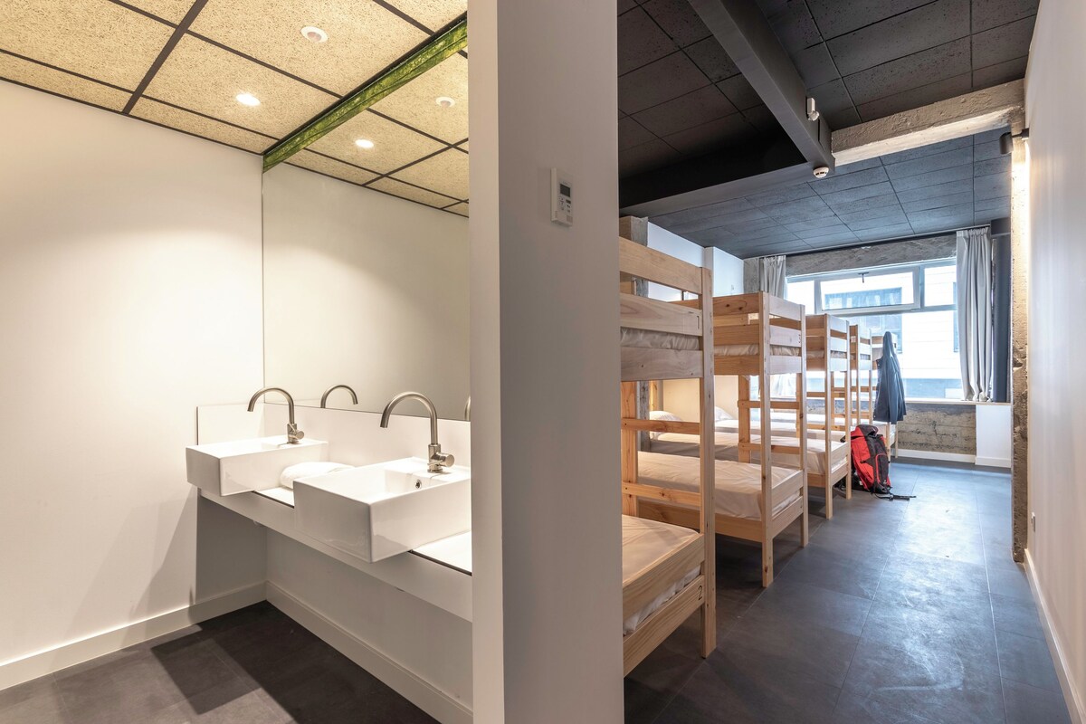 A modern bathroom with two sinks and a large mirror. There are bunk beds in the adjacent room, and a window with curtains.