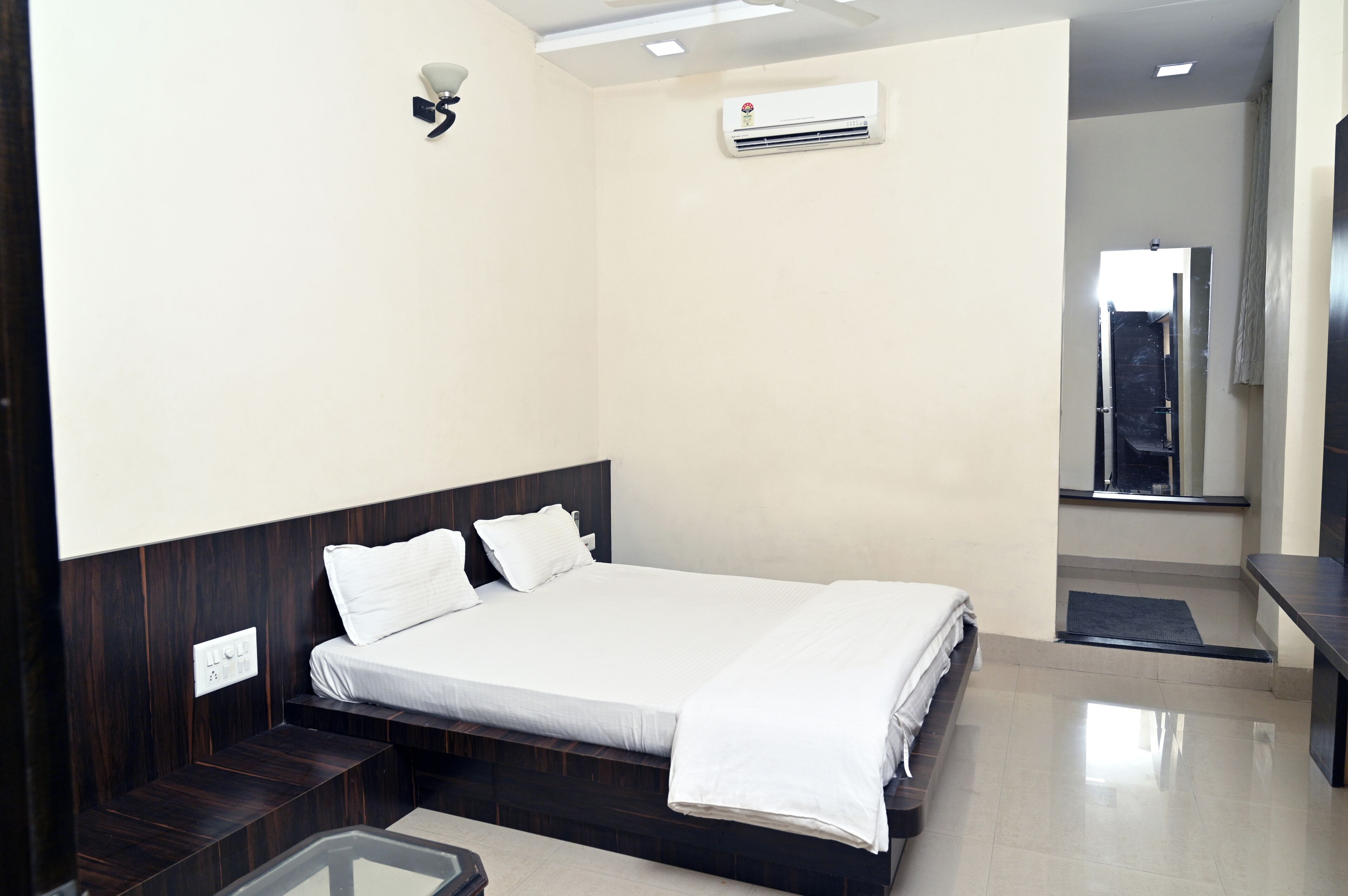 A modern hotel room with a large bed, a small seating area, a mirror, and an air conditioning unit.