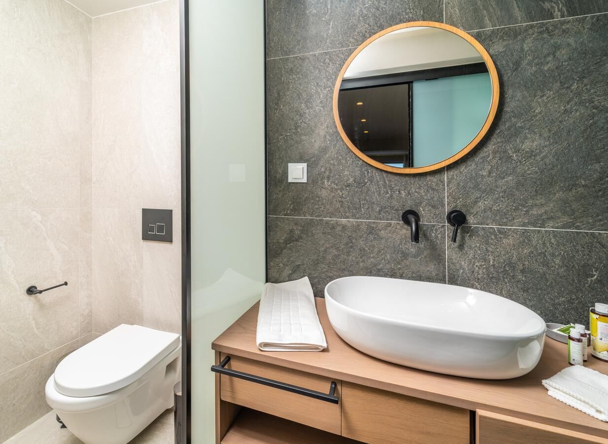 A modern bathroom with a toilet, a large oval sink, a wooden vanity, a round mirror, and a glass shower enclosure.