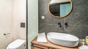 A modern bathroom with a toilet, a large oval sink, a wooden vanity, a round mirror, and a glass shower enclosure.