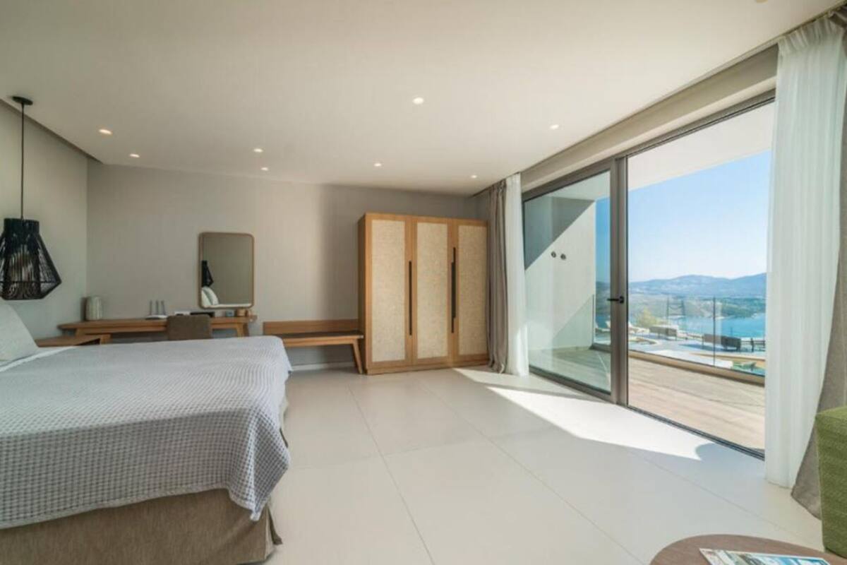 A modern hotel room with a bed, a desk, a chair, and a wardrobe. There is a large window with a view of the sea and mountains.