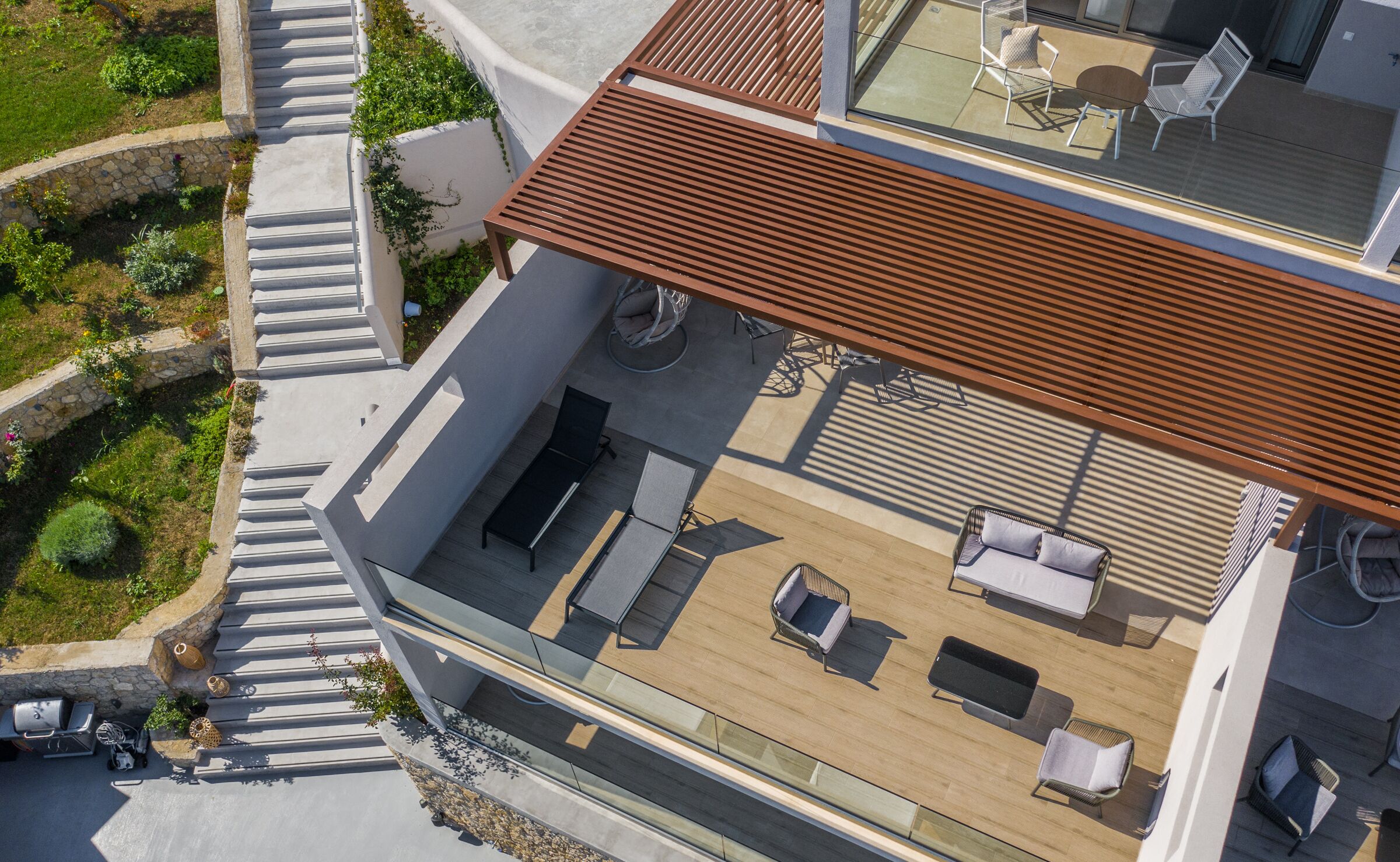 A modern outdoor deck with a wooden floor, seating area, and a glass railing overlooking a garden.