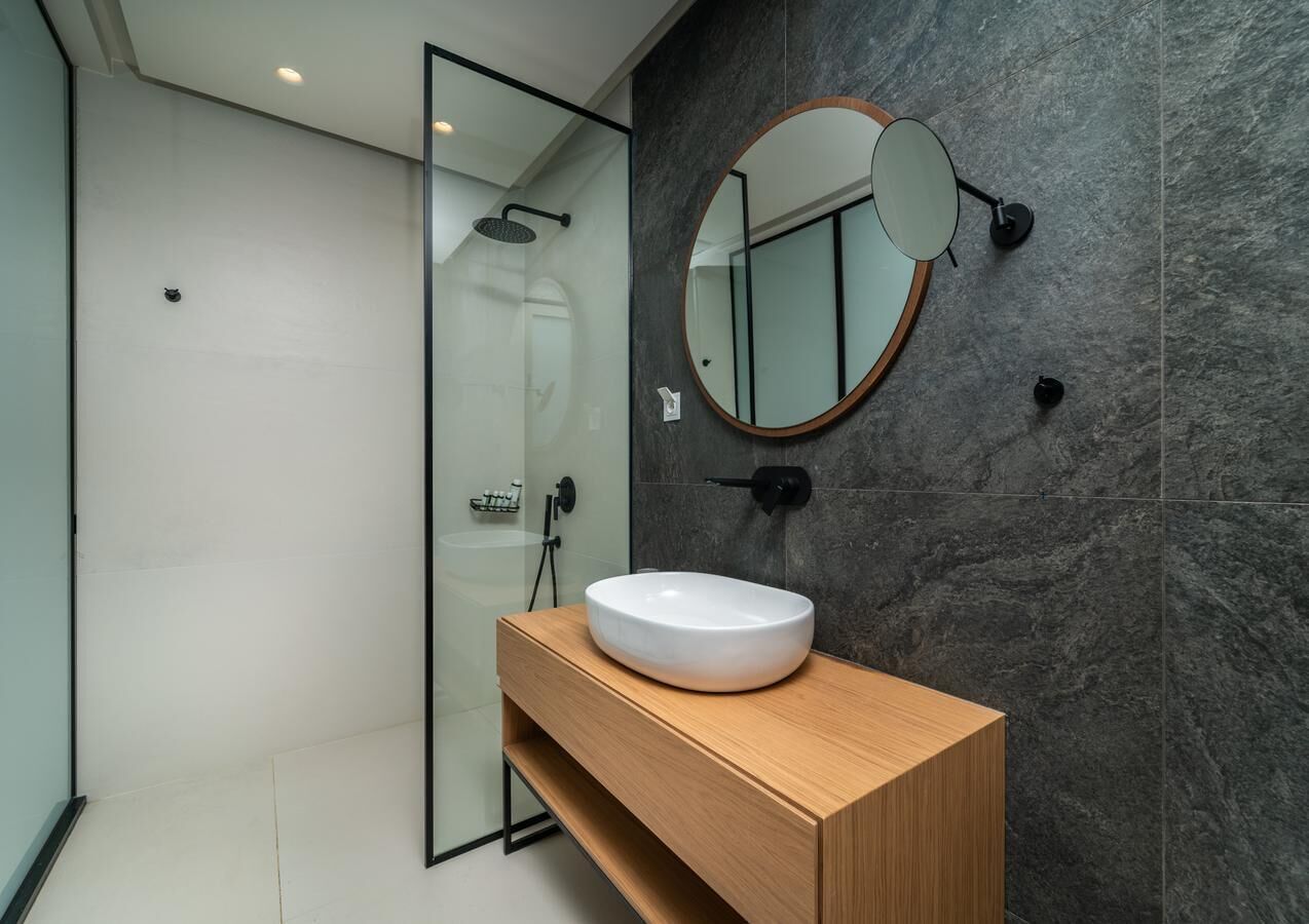 Un baño moderno con cabina de ducha de vidrio, tocador de madera con lavamanos blanco y un espejo redondo en la pared.