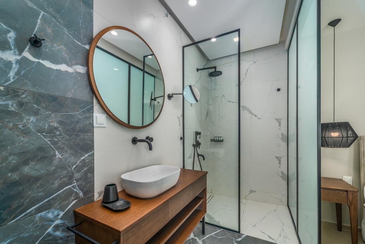 Une salle de bain moderne avec un sol en marbre, un meuble-lavabo en bois doté d’un vasque, un miroir rond et une cabine de douche en verre.