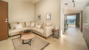 A modern living room with a beige sofa, a coffee table, and a patterned rug. There are framed pictures on the wall and a view of the outdoors through a window. - Katouna Suites Luxury Boutique Hotel - Adults Only (Lefkada)