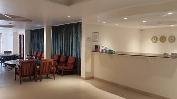 Zona de recepción de hotel con mostrador, área de estar con sillas y una mesa, y un gran ventanal con cortinas.