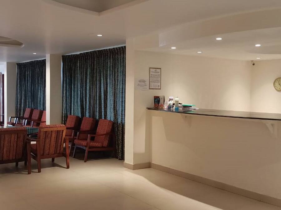 Zona de recepción de hotel con mostrador, área de estar con sillas y una mesa, y un gran ventanal con cortinas.