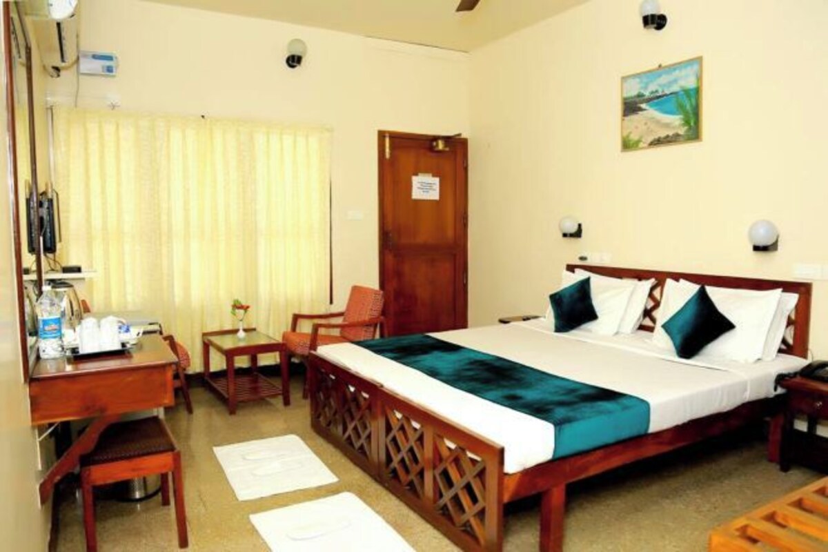Habitación de hotel con una cama de madera, un escritorio con silla, una mesita y un ventanal con cortinas.