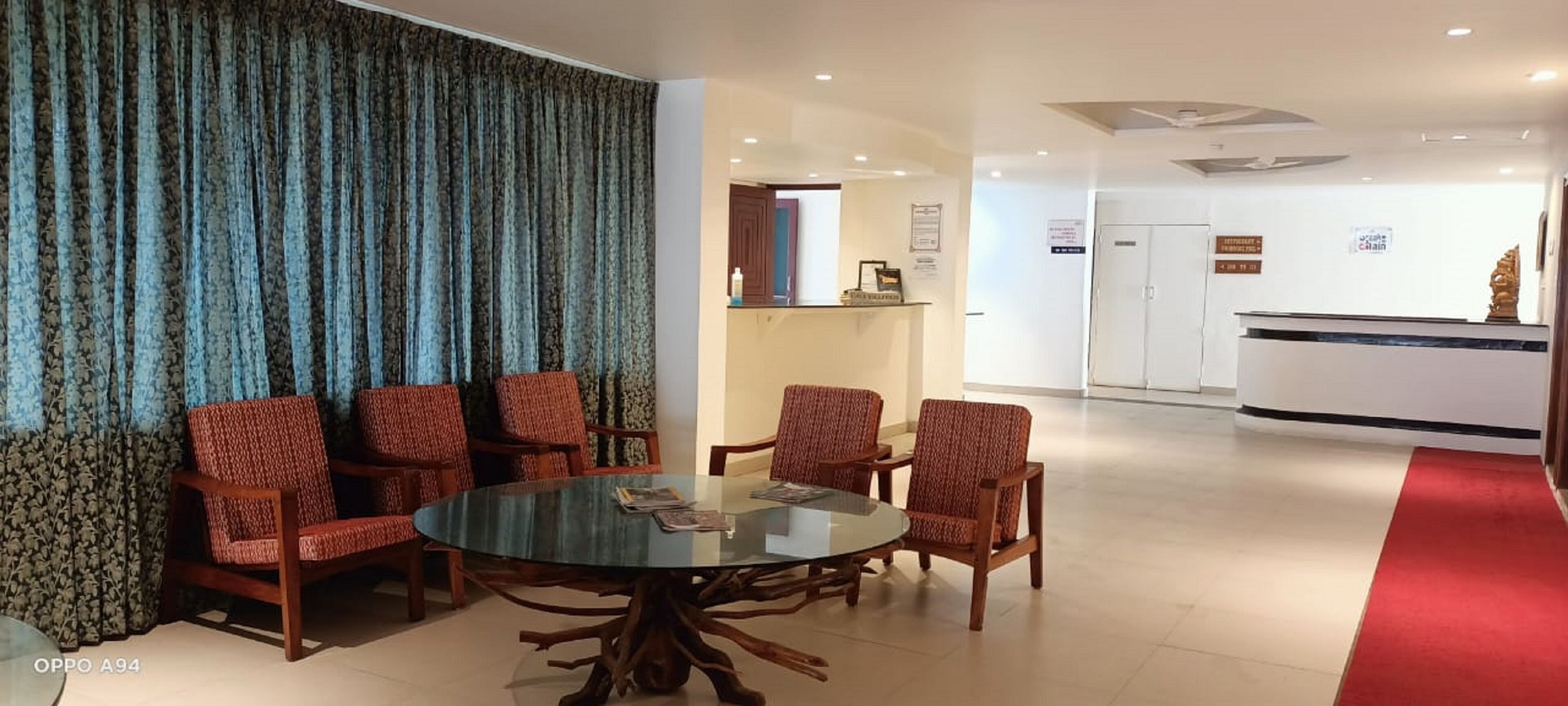 A hotel lobby with a round glass table surrounded by six patterned chairs, a reception desk, and a red carpet leading to the entrance.