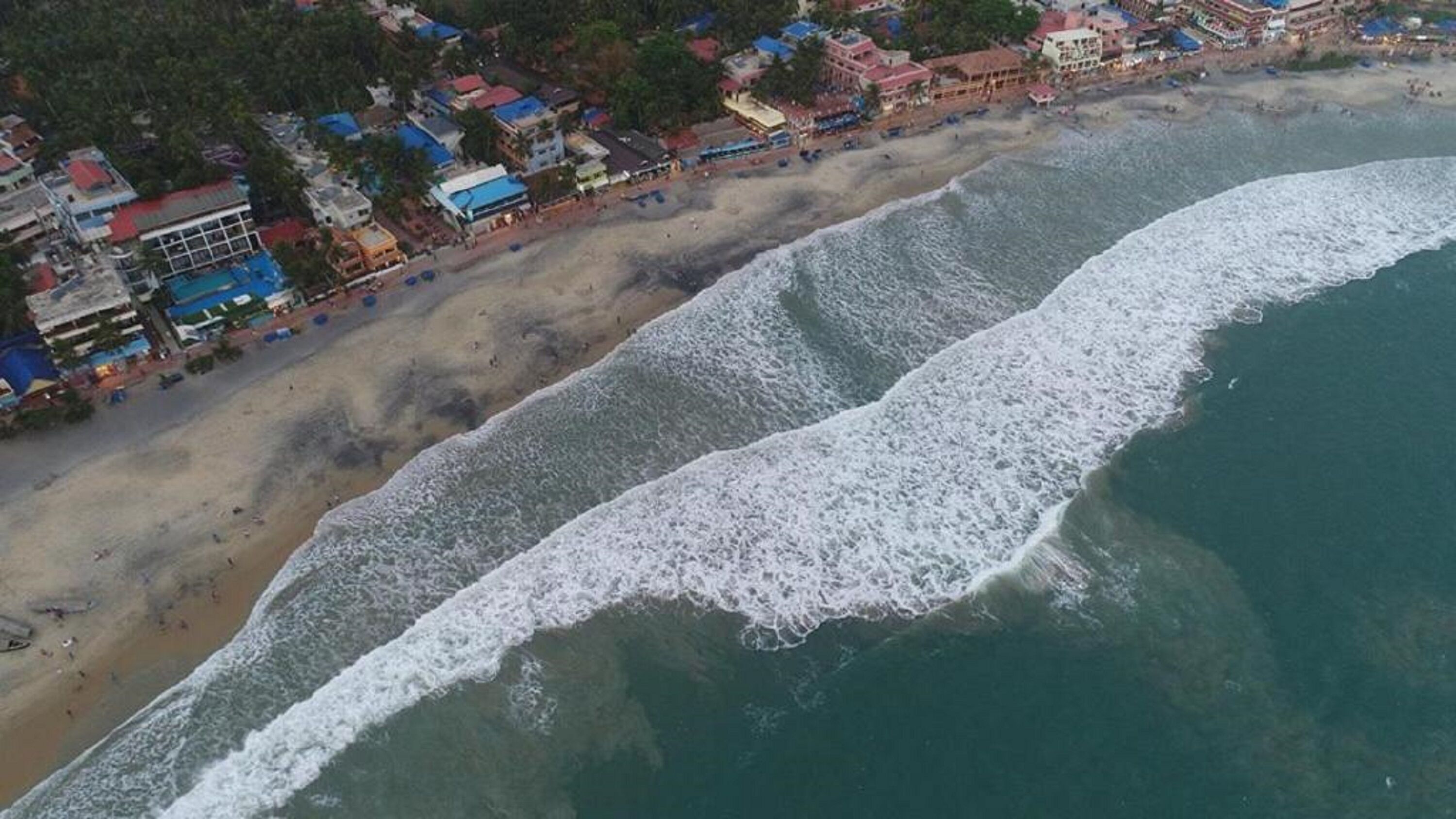 Ovenudblik over en strand med en lang kystlinje, bygninger langs kysten og bølger der nærmer sig land.