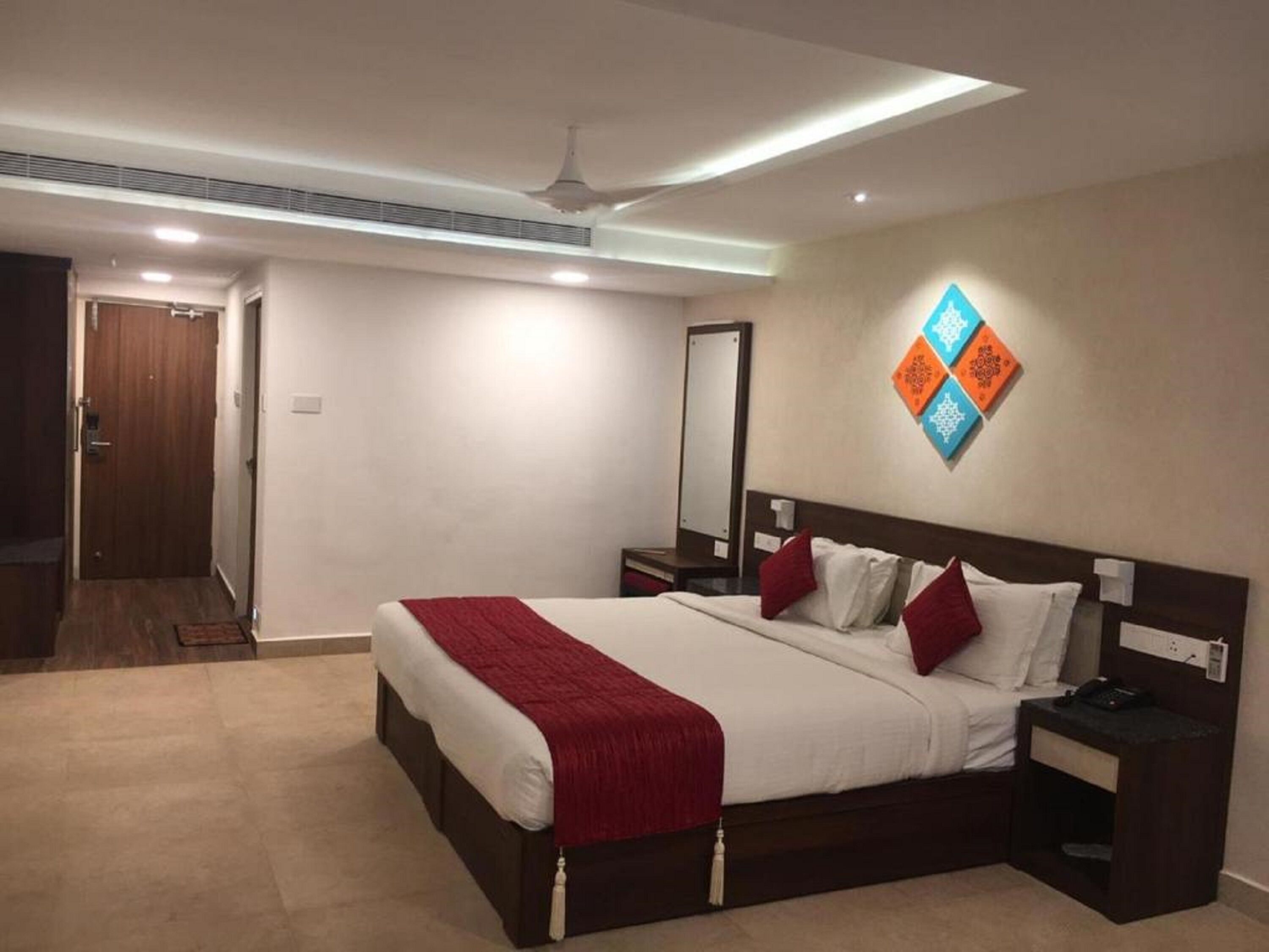 Una habitación de hotel con una cama grande, una mesita de noche, un ventilador de techo y un espejo montado en la pared.