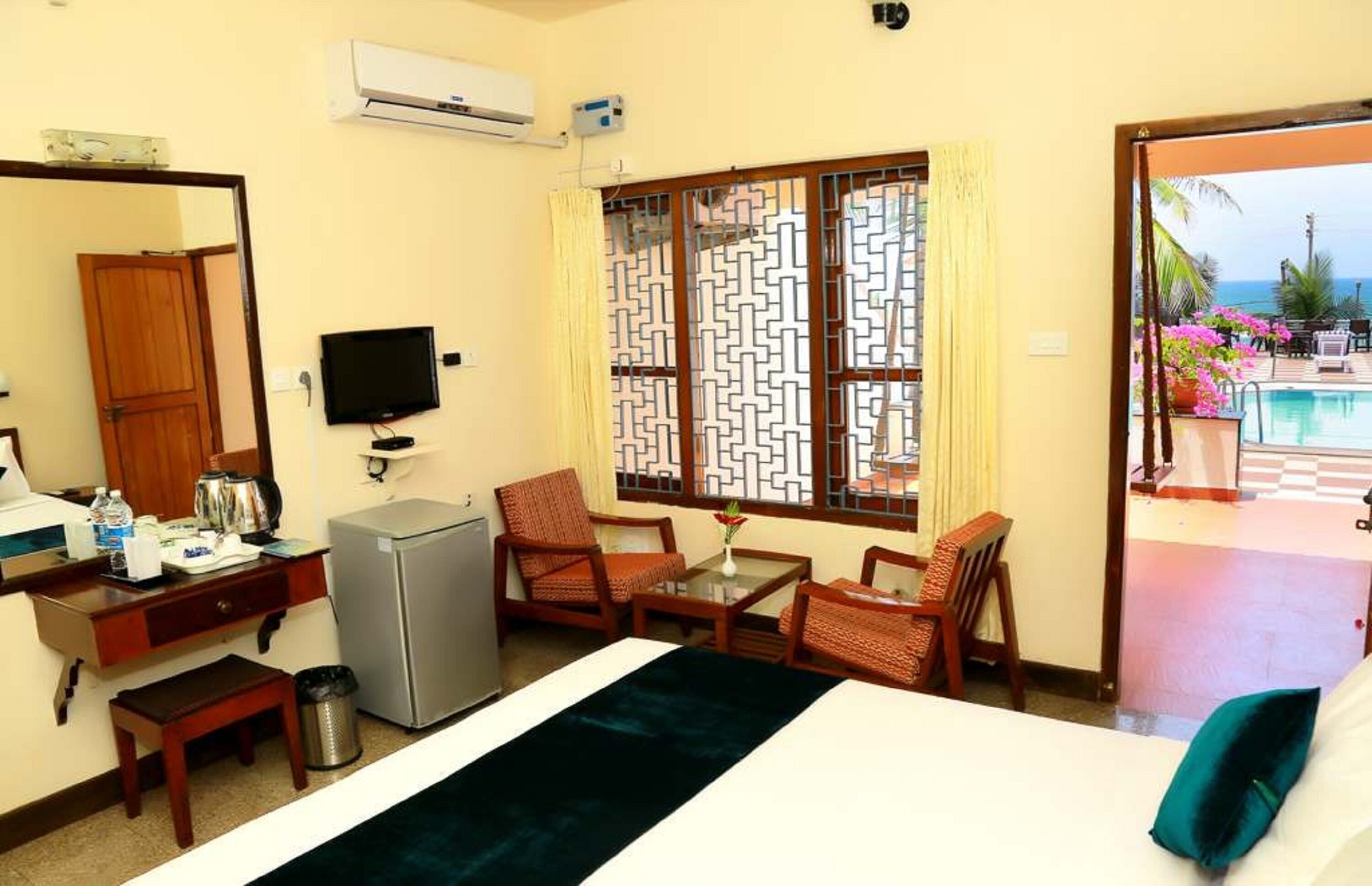 A hotel room with a bed, a desk with a computer, a TV, and a window with a view of a pool and the sea.