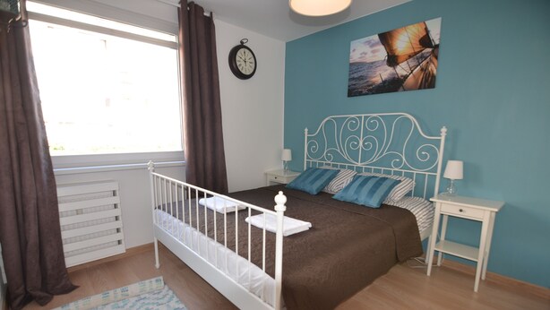 A bedroom with a white metal bed frame, a brown bedspread, a blue wall, a clock, and a window with curtains. - Victus Apartamenty - Marina (Sopot)