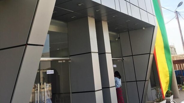 Un bâtiment moderne à la façade géométrique, doté d’une entrée en verre et d’un mât sur lequel flotte un drapeau.