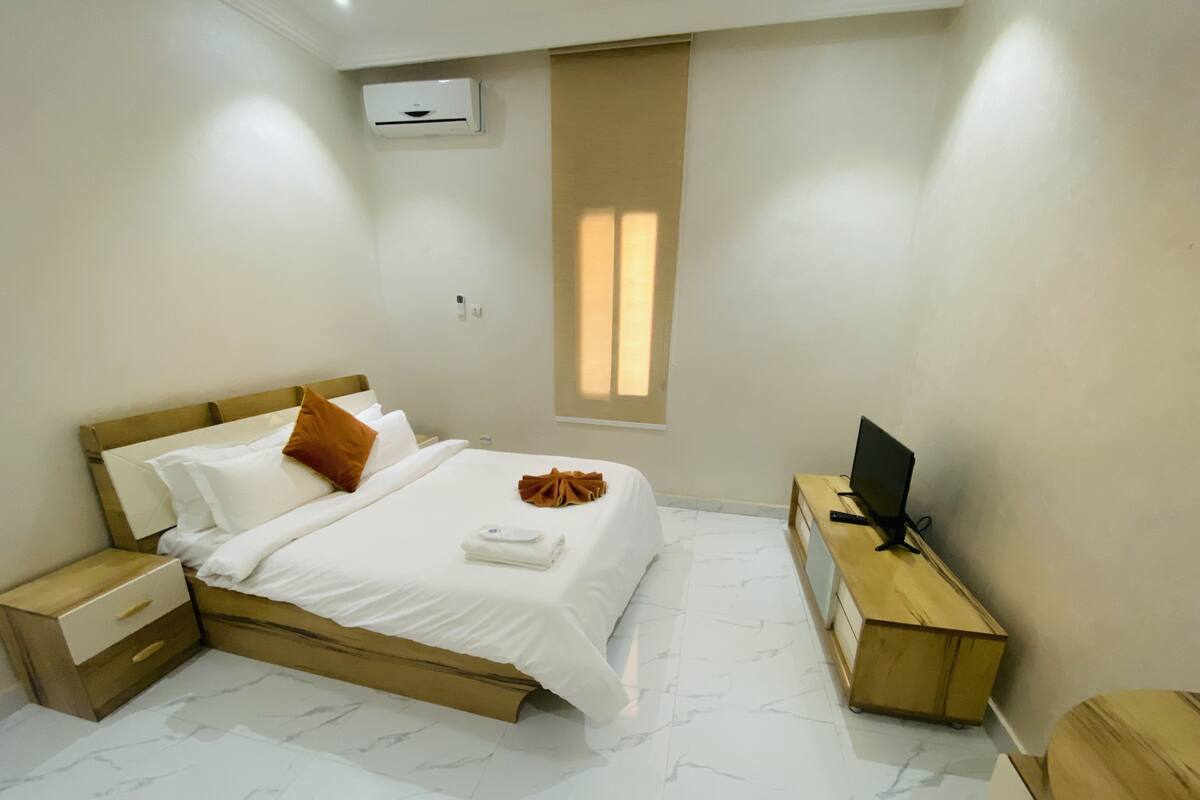 A bedroom with a bed, a TV, a nightstand, and a window.