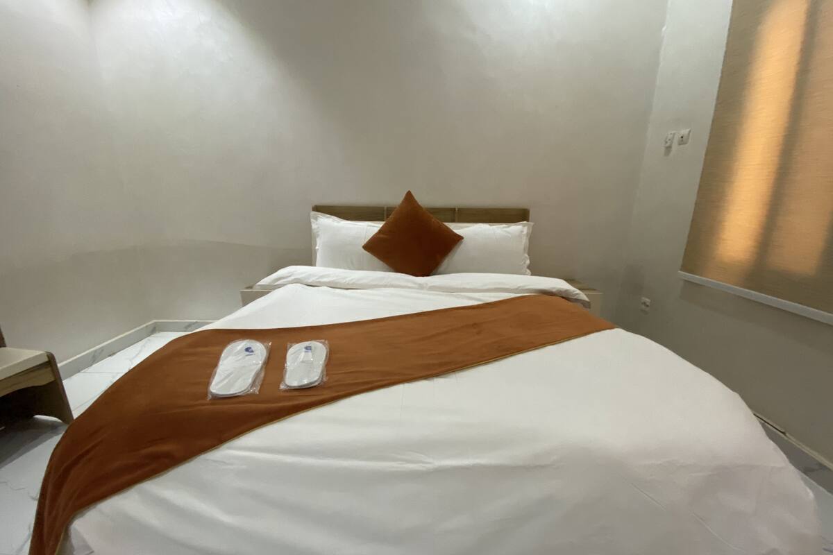A hotel room with a bed, white linens, a brown throw blanket, and two slippers on the bed.
