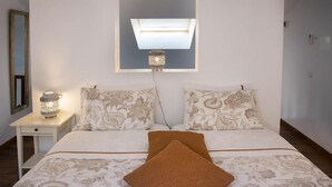 Un dormitorio con cama, dos almohadas, una lámpara de noche, un espejo y un tragaluz sobre la cama.