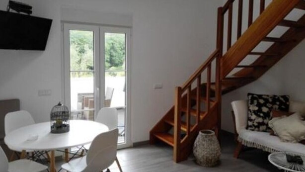 Una sala de estar con una mesa redonda blanca, un sofá con cojines estampados, una escalera de madera y un amplio ventanal con vistas a la vegetación.
