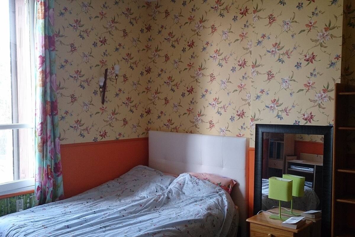 A bedroom with a single bed, floral wallpaper, a window with curtains, and a nightstand with a lamp.